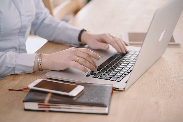 Woman's hands typing on laptop. Next to laptop is a closed notebook with a cellphone on top.