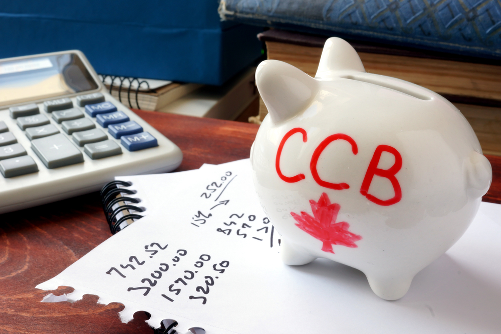 Piggy bank with CCB (Canada Child Benefit) written on it and standing on papers with numbers hand-written on them next to a calculator