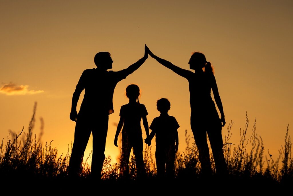 Family of four stands in a field during sunset. The parents on each side form a "roof" shape with their arms over their two children.