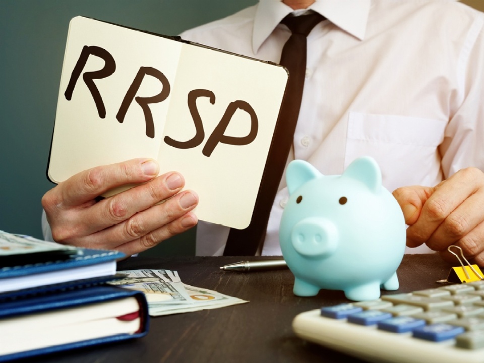 Man holding a sign with RRSP written on it with black marker. Man sitting behind desk. All we can see is his white shirt and black tie, his hand with the sign, a blue piggy bank on his desk, a corner of an adding machine and some notebooks.