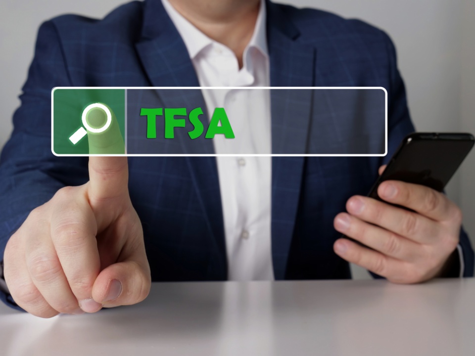 Finger pressing on a magnifying glass icon saying "TFSA", In the background we can see a torso of a person wearing a white shirt and a dark suit jacket. We can't see a face.