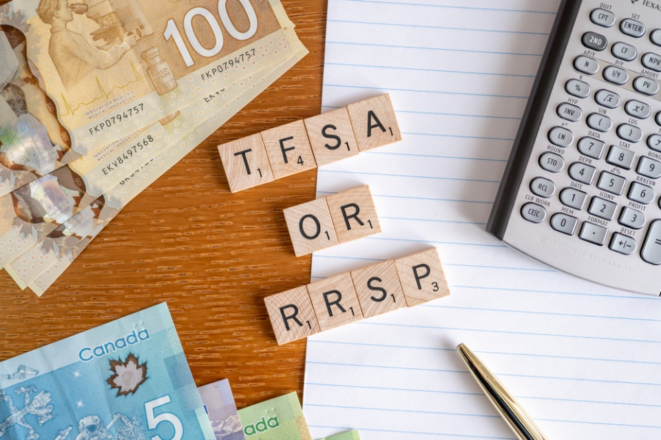 "TFSA or RRSP" on a Scrabble blocks lying on top of a lined blank notebook next to a pencil and a calculator and next to some Canadian paper money - some hundred dollar bills and five dollar bills.