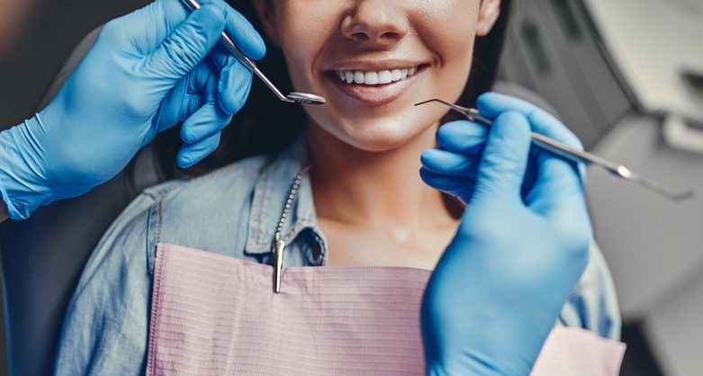 Young woman in stomatology clinic smiling. Bottom of face visible. Gloved dentist hands holding dental implements.