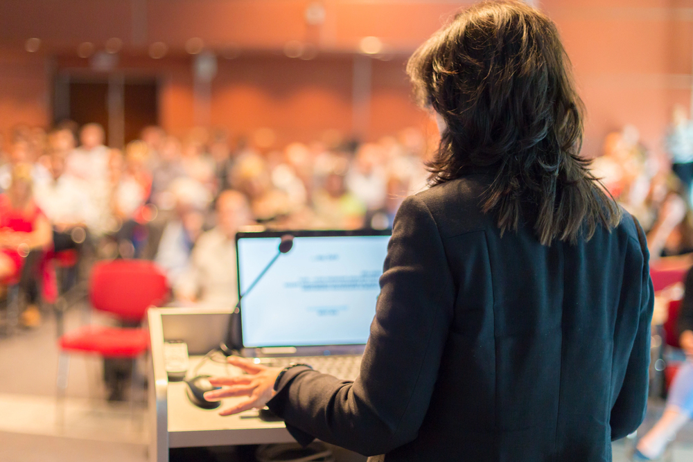 person with long hair with their back to the camera, speaking to a microphone in front of a blurry audience