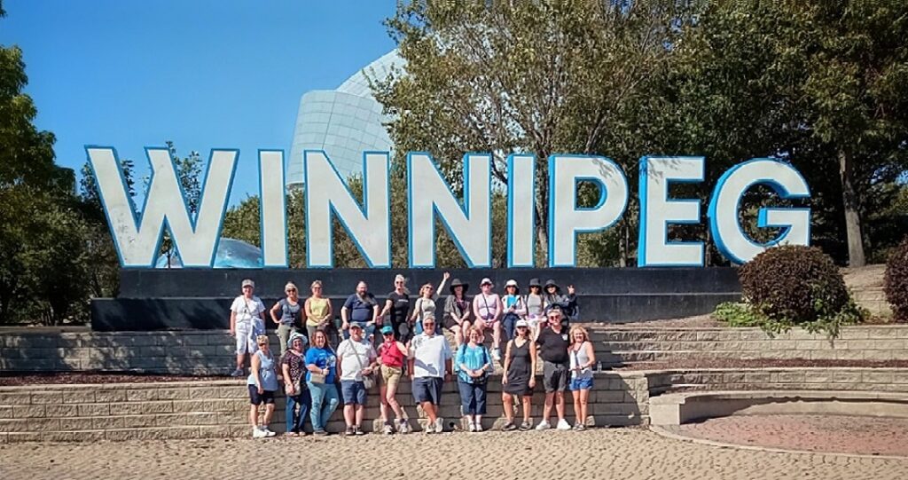 Group of about 20 people in front of the Winnipeg city sign at the Forks, Human Rigths in background, group led by Bea Dabrowski