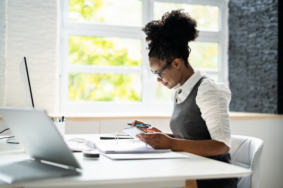 woman with curly hair and glasses smiling and sitting at a desk smiling and looking down at documentation