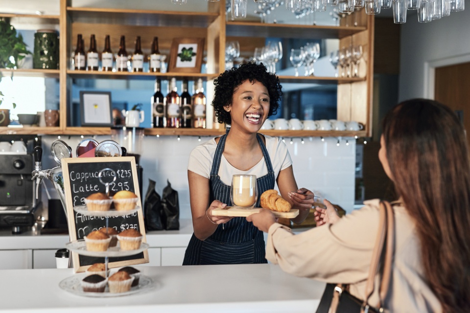 smiling barista passes a coffee and a plate with a baked good to a female customer