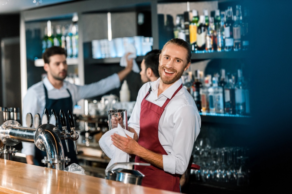 smiling barman in a red apron looks directly at camera while wiping down a glass. His colleague in the background is blurry.