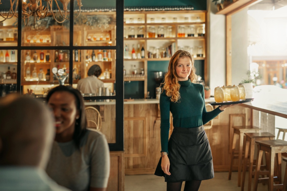 waitress with long reddish hair wearing green sweater and black skirt is carrying a tray with drinks to a table in a pleasantly lit restaurant. Smiling patron woman in foreground.